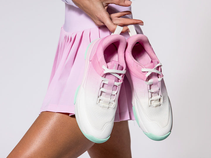 Pair of women’s Court2 pink and white pickleball shoes with mint soles held by model in pleated pink skirt