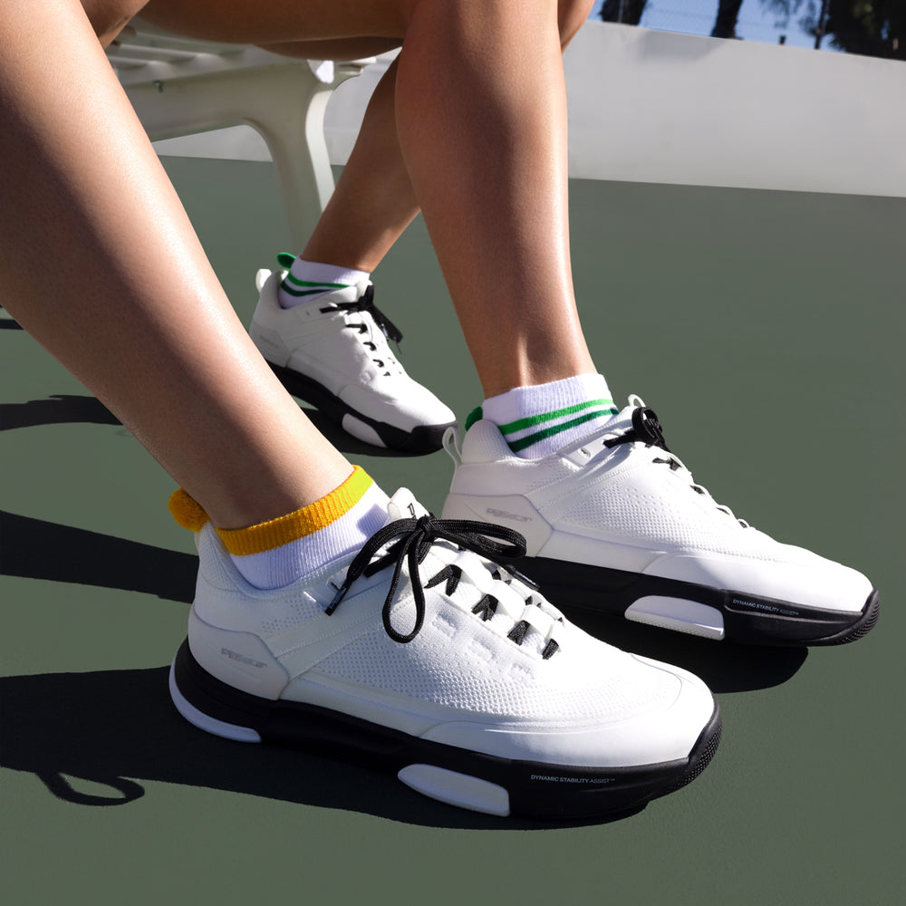Close-up of two people wearing white PB5 court sneakers on an outdoor tennis/pickleball court.