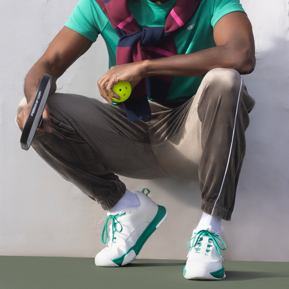 Person crouching courtside holding a paddle and ball, wearing white court sneakers with jade-green accents.