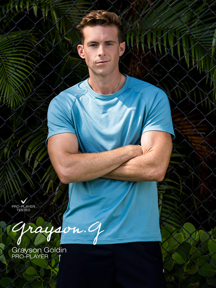 Pro pickleball player Grayson Goldin wearing Arctic Core Performance Tee and black Signature Court Shorts standing in front of a fence with greenery background