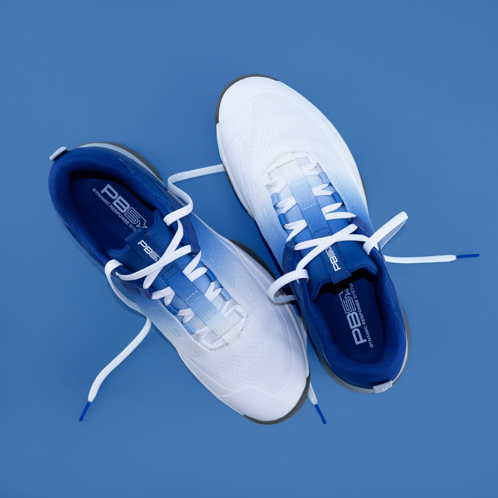Flat-lay product shot of white-and-navy PB5 sneakers with loose laces on a blue backdrop.