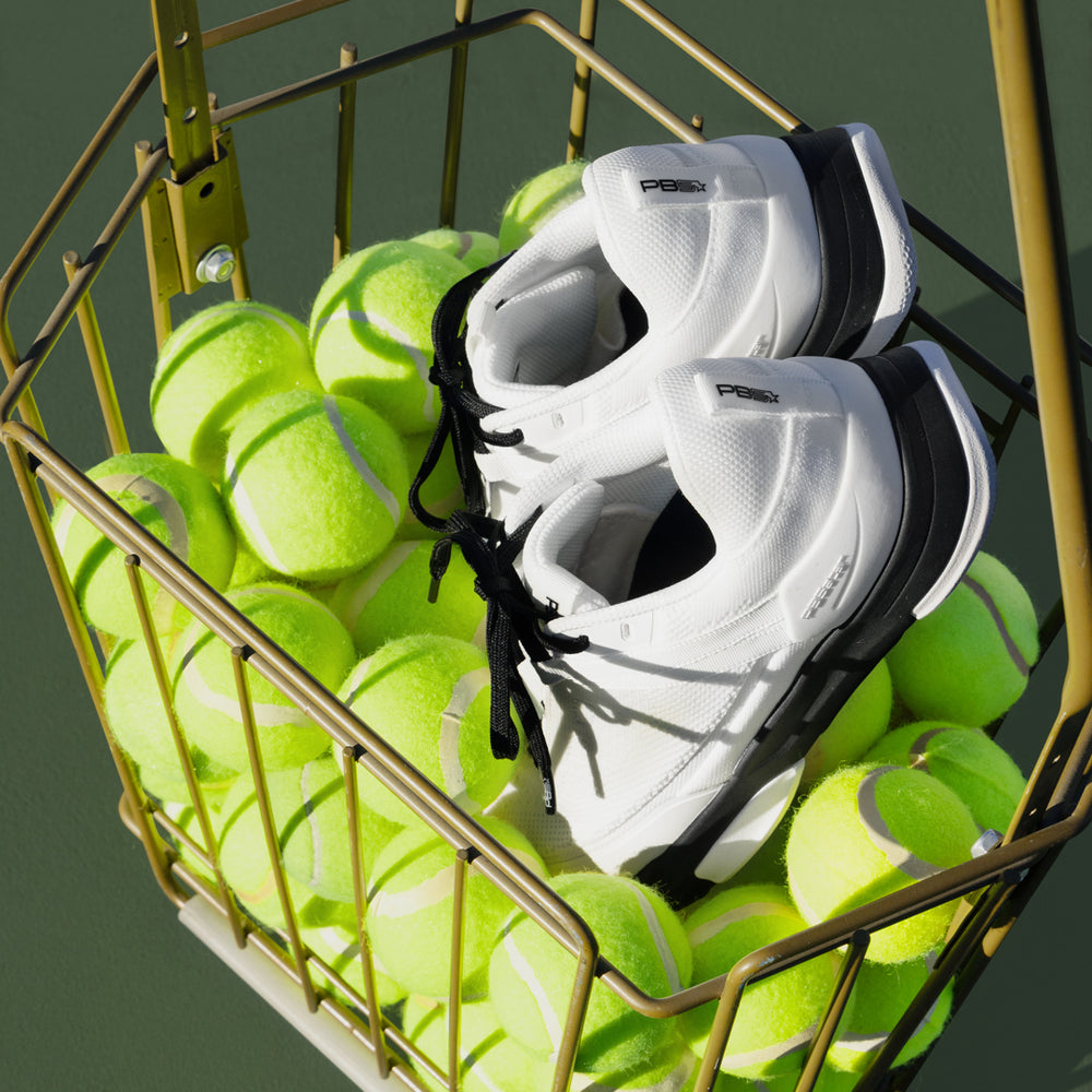 Pair of white court sneakers with black details sitting on top of bright tennis balls in a metal basket.