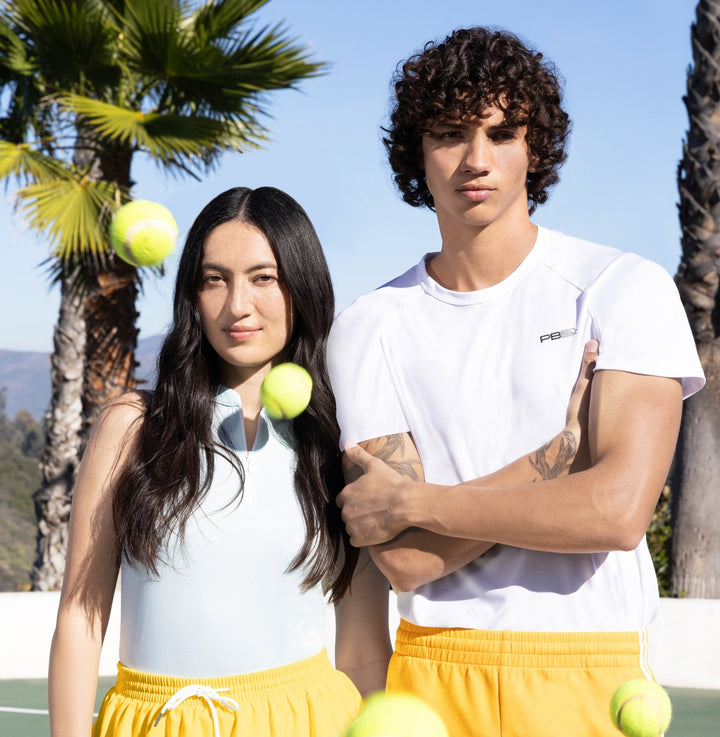 Models wearing a white PB5 athletic shirt in an outdoor tennis lifestyle shot with tennis balls