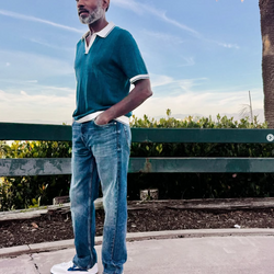 Man wearing a teal polo shirt and blue jeans standing outdoors with a palm tree in the background wearing PB5 Cosmic shoes in white-blue.