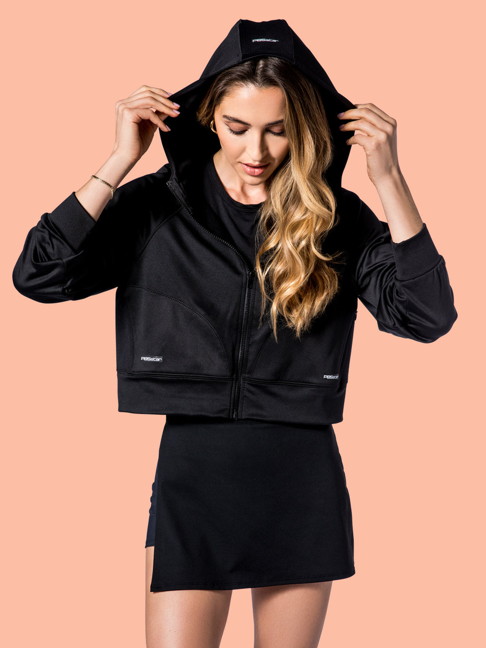 Model adjusts the hood of PB5Star's black Cropped Performance Hoodie, pairing it with a black Side Split Skirt for a sporty-chic look.